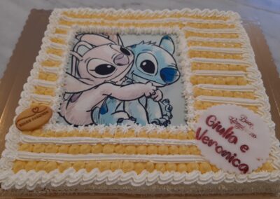 Due Gu incomparable e teneri in un cake decorato con panna e palline di crema, con immagine di due simpatici personaggi animati al centro, per occasioni speciali come battesimi o compleanni.