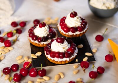 Delicati crostatine alla crema con ciliegie fresche decorano un vassoio, circondate da mandorle e ciliegie sparse. Una dolce opera che unisce sapori autentici e presentazione invitante, perfetta per ogni occasione speciale.