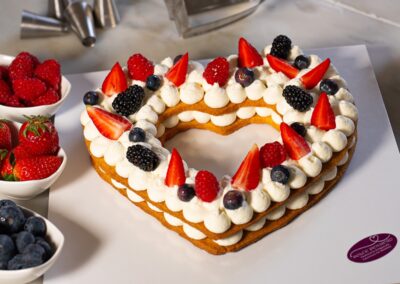 Deliziosa torta a forma di cuore decorata con panna, fragole, lamponi, mirtilli e more, perfetta per occasioni speciali o festeggiamenti. La presentazione elegante valorizza ogni morso.
