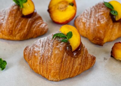 Croissants farciti con pesca e menta, decorati con zucchero a velo e presenti su un piano in carta velina, perfetti per colazioni e brunch. Dolce Pensiero Pasticceria offre dolci artigianali di alta qualità.
