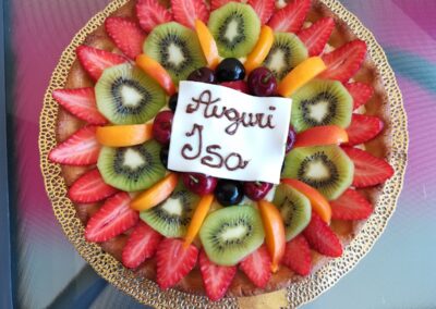 Fragola, kiwi, mirtilli e pesche su torta decorata con frase "Auguri Isa" in cioccolato bianco. Pasticceria fresca e colorata, ideale per occasioni speciali.