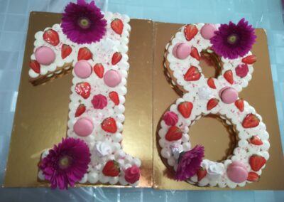 Torta a forma di numero 18 decorata con panna, fragole, macaron rosa e fiori viola, perfetta per celebrazioni speciali e compleanni.