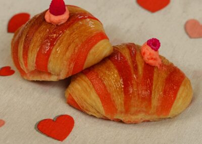 Croissant decorati con strisce di arancia e guarniti con lamponi, ideale per dolci romantici e colazioni golose, foto su sfondo di cuori rossi e rosa.
